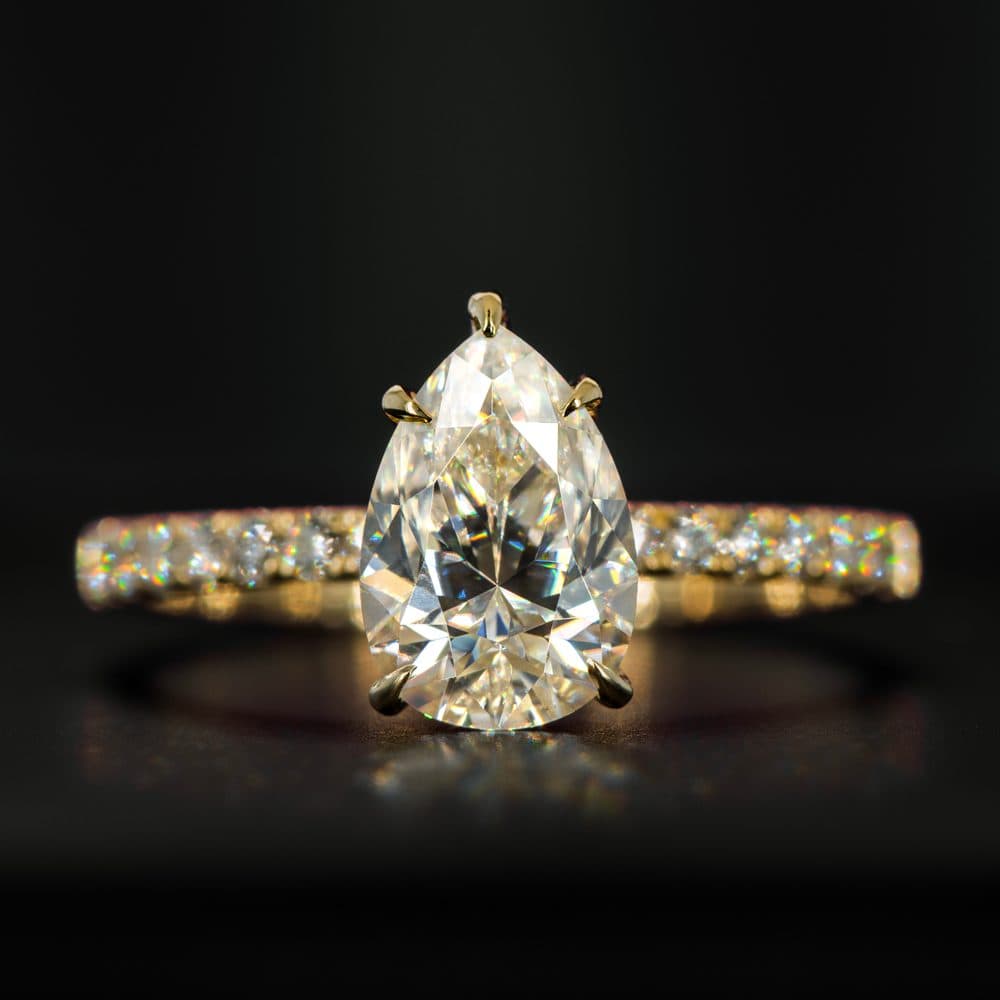 A yellow gold pear-cut diamond solitaire engagement ring with a pavé band on a dark background