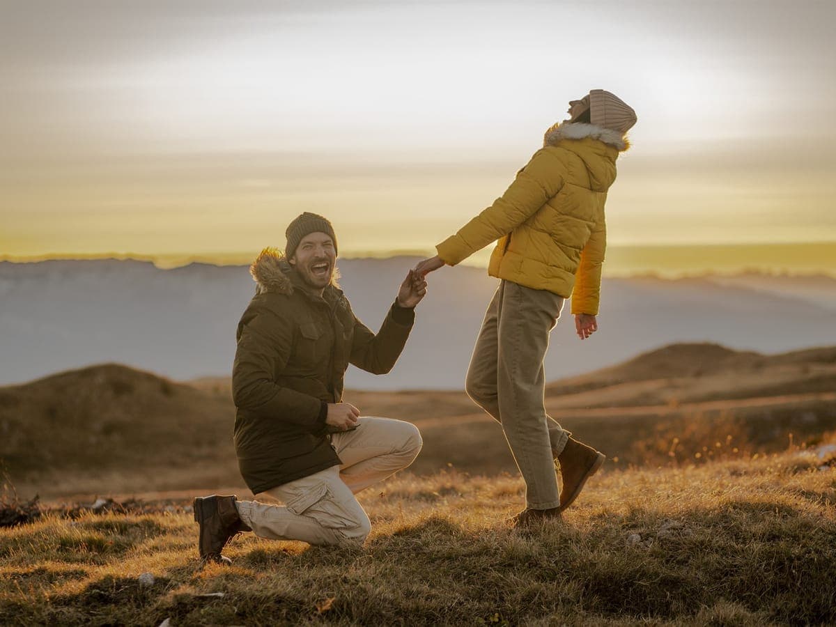 A couple on a coastal clifftop at sunset, one down on one knee proposing