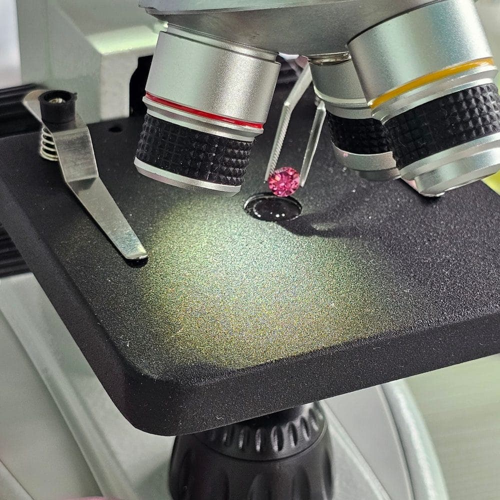 A loose lab-grown diamond held in tweezers beneath a laboratory microscope, test tubes blurred behind