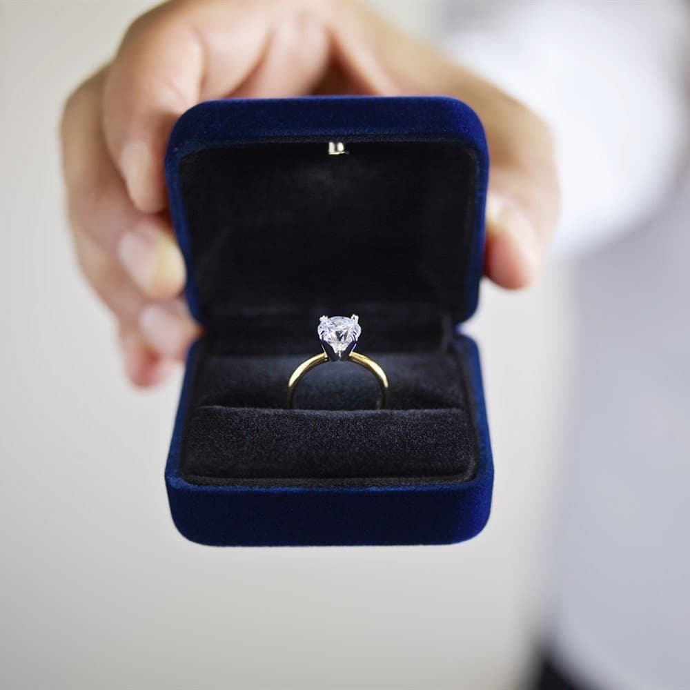 Two hands holding open a navy ring box, presenting a diamond solitaire engagement ring